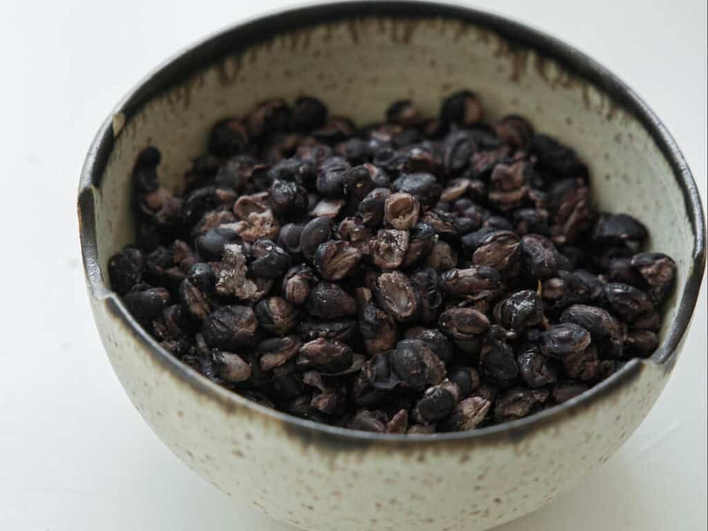 A ceramic bowl filled with black beans rests on a white surface. The beans appear cooked or roasted, and the bowl has a speckled, rustic design.